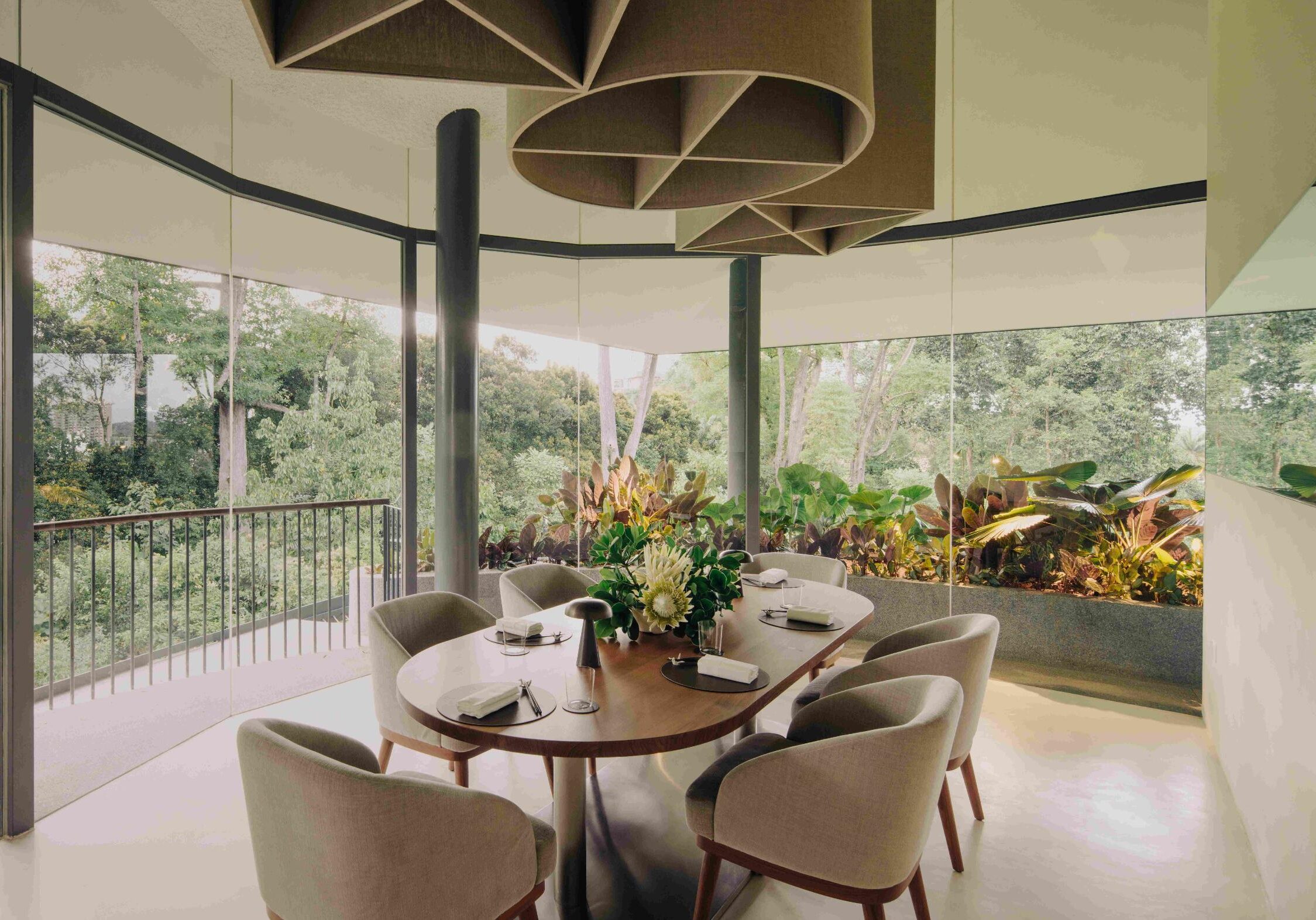 This modern, minimalist dining room features a curved wooden table surrounded by plush grey chairs, all set against floor-to-ceiling windows that offer a panoramic view of lush green foliage. Unique, geometric acoustic ceiling fixtures hang above the table, which is elegantly set with a floral centerpiece and sleek tableware.