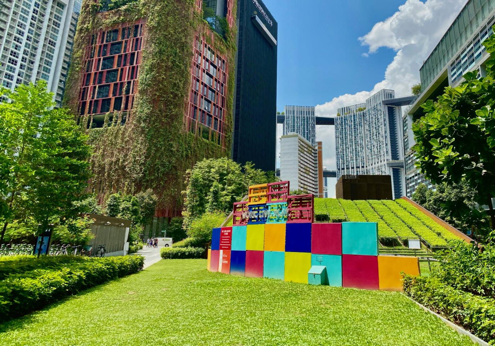 This vibrant urban landscape features a striking high-rise building covered in vertical gardens rising above a lush, sunny park. In the foreground, a playful installation of bright, multicolored cubes sits on a manicured lawn, emphasizing the harmonious blend of modern architecture and green space.