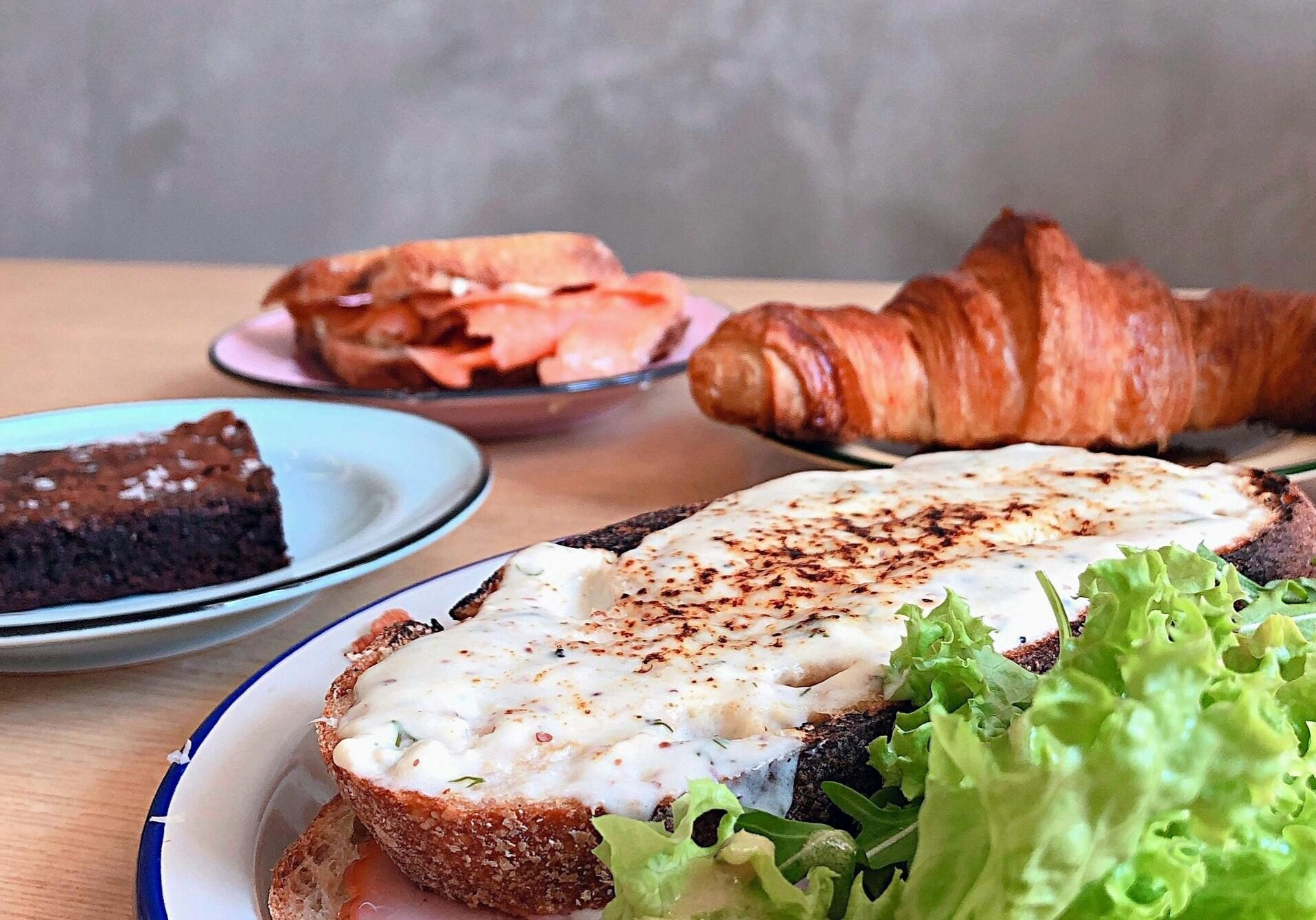 An inviting brunch spread is arranged on a wooden table, featuring a prominent slice of rustic toast topped with a creamy melted topping and fresh greens. Surrounding the main dish are a golden croissant, a smoked salmon sandwich on a pink plate, and a rich chocolate brownie, offering a variety of sweet and savory treats.