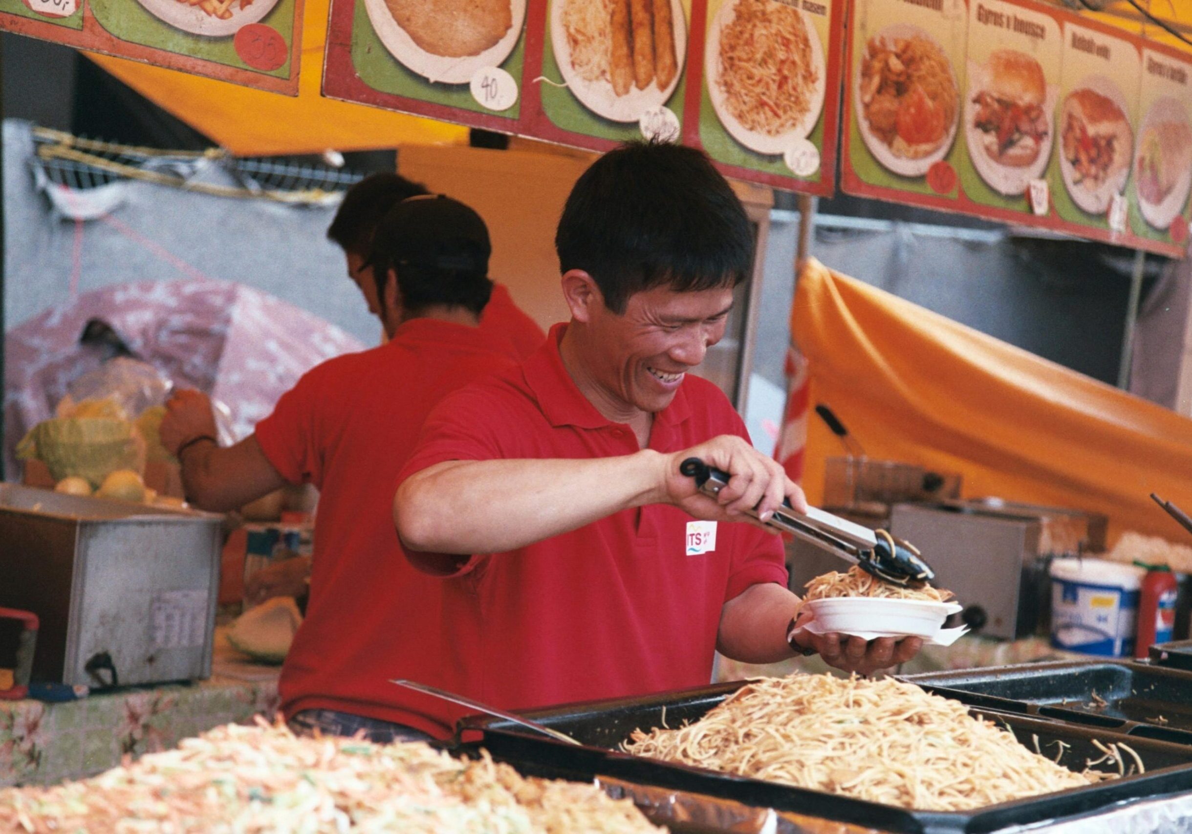 A smiling vendor in a red polo shirt uses tongs to serve a portion of noodles from a large tray into a white bowl. The scene captures a bustling street food stall with overhead menu pictures and another worker busy in the background.