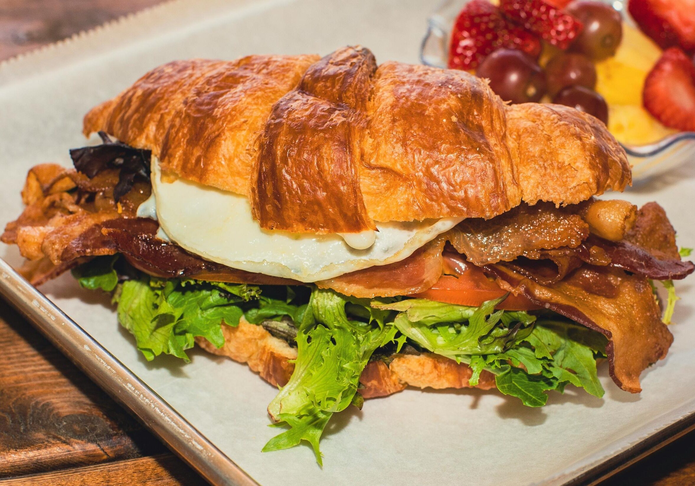 A golden, flaky croissant sandwich is piled high with crispy bacon, a fried egg, and fresh green lettuce, sitting on a sheet of parchment paper. In the background, a small glass bowl holds a refreshing side of fruit, including red strawberries and grapes.