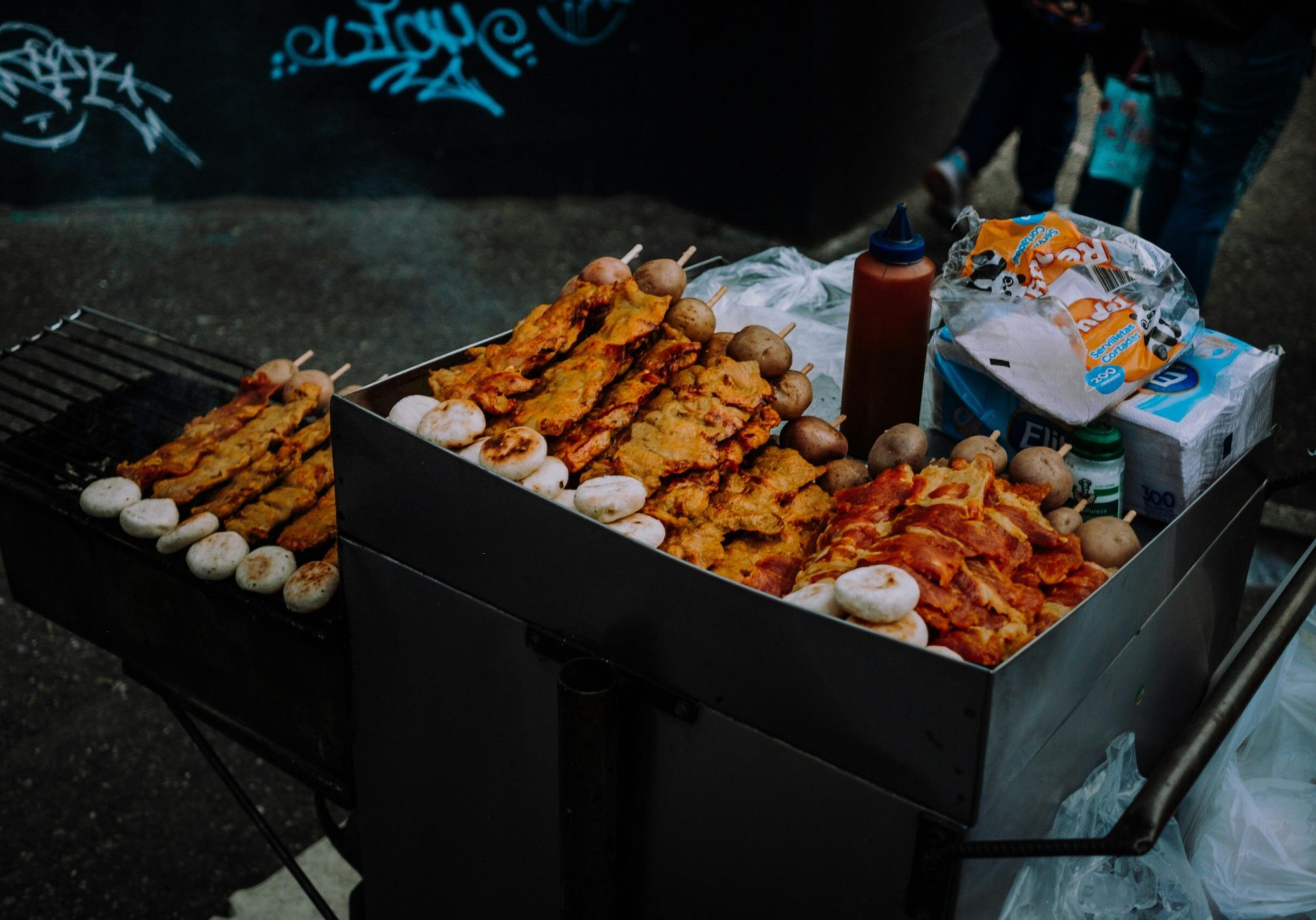 A street food cart is densely packed with grilled meat skewers and round buns, with additional items cooking on a smaller grill to the left. The setup includes a squeeze bottle of sauce and napkins, situated against a dark wall tagged with blue graffiti.