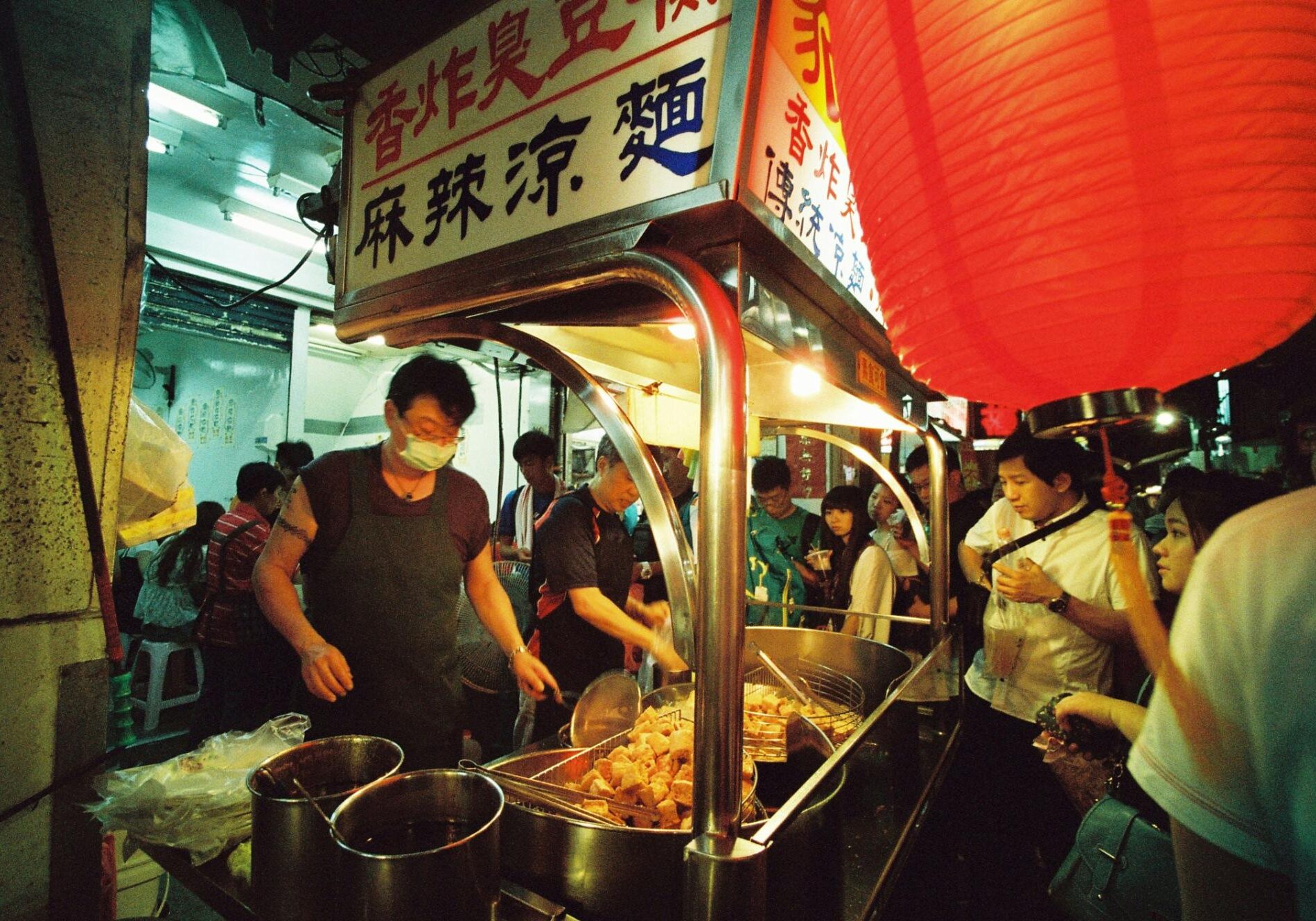 Beneath the warm glow of a large red lantern, a masked vendor in an apron tends to a busy food stall at a night market. Large metal vats filled with golden fried snacks sit at the counter as a crowd gathers near the illuminated signage featuring Chinese characters.