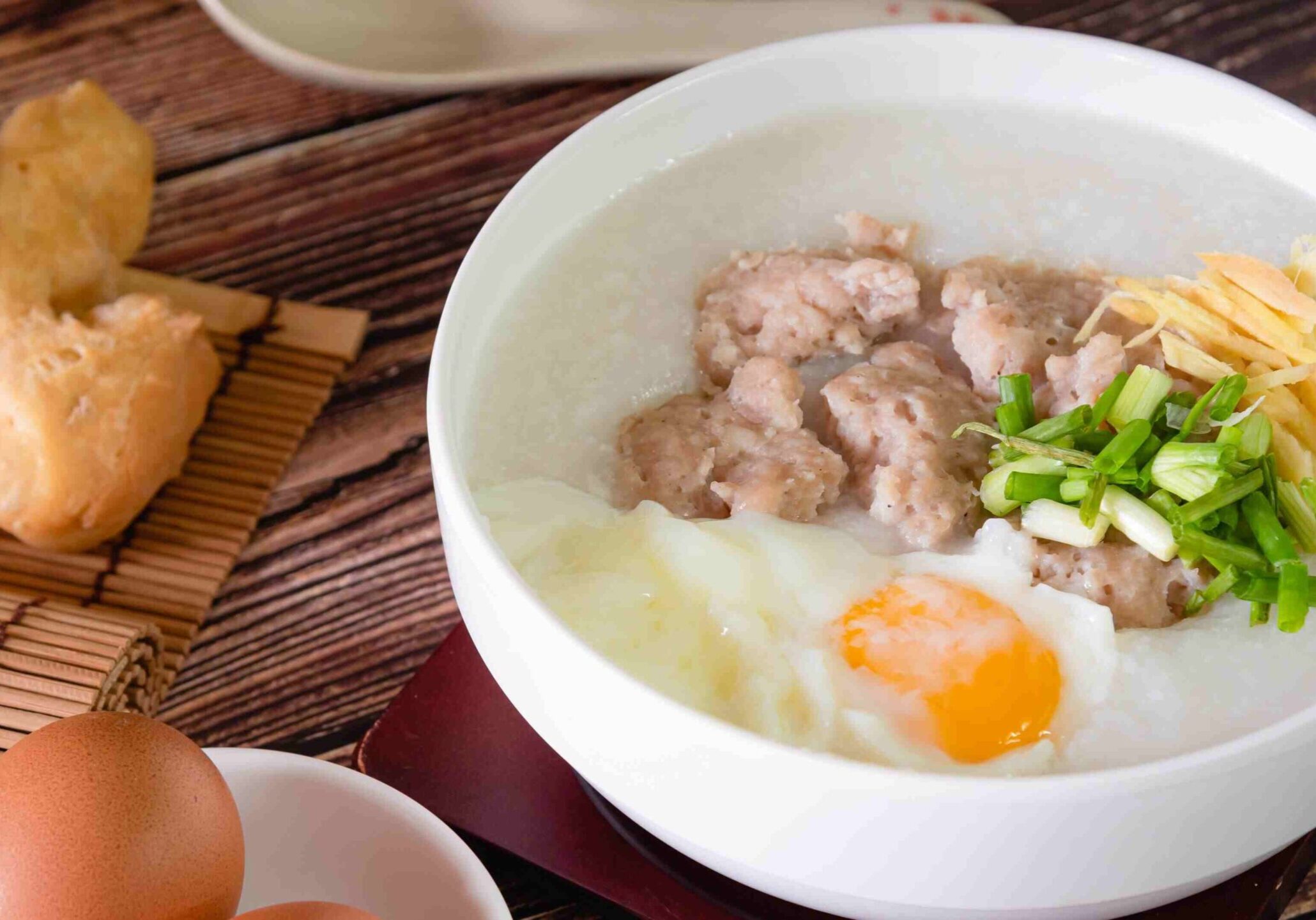 A white bowl of creamy Chinese congee is topped with savory pork meatballs, a soft-poached egg, fresh ginger slivers, and chopped green onions.