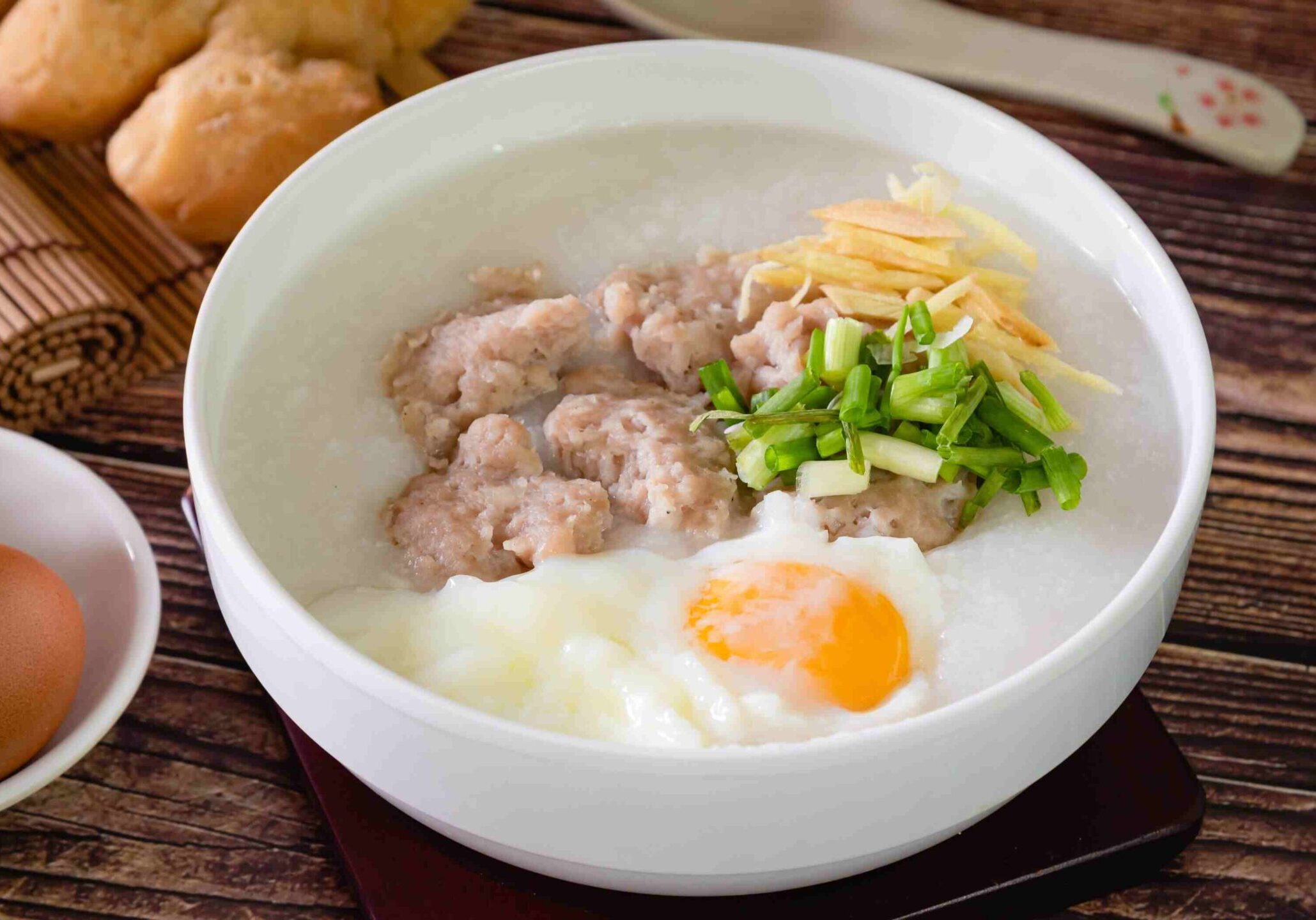 This image shows a white bowl filled with Chinese rice porridge, or congee, topped with seasoned pork meatballs, a soft-poached egg, sliced ginger, and green onions.