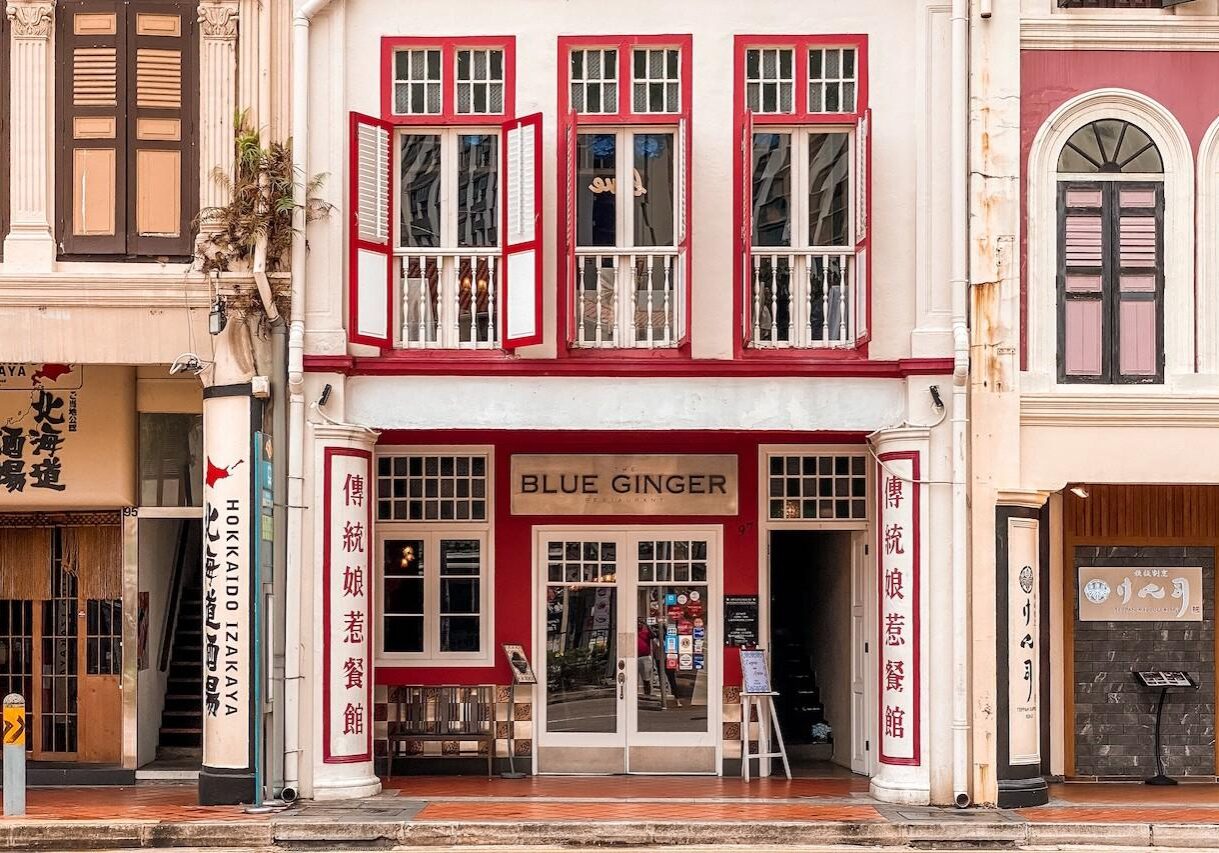 The storefront of The Blue Ginger Restaurant is housed in a classic white shophouse with vibrant red accents around the windows and entryway. The ground floor features white pillars with vertical Chinese calligraphy, while the upper floor displays traditional wooden shutters painted in matching red and white.