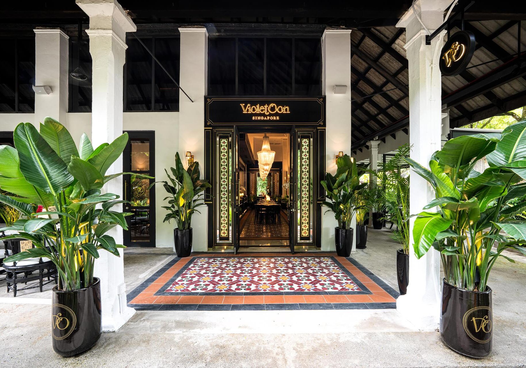 The entrance of Violet Oon Singapore features a striking black doorway framed by intricate Peranakan-style tiles and lush, oversized potted plants. Large white columns support an open-air veranda, while a glimpse through the open doors reveals a warm, elegant interior lit by grand chandeliers.