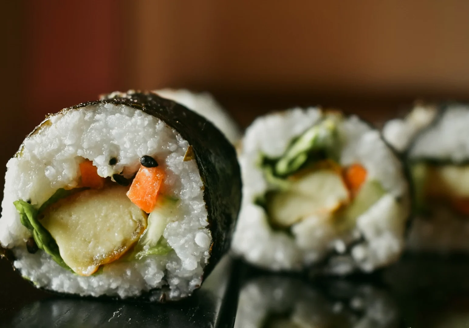 Macro close-up, eye-level shot of a sliced vegetable sushi roll wrapped in nori, showing white rice, avocado, carrot, and cucumber with shallow depth of field on a dark reflective surface.