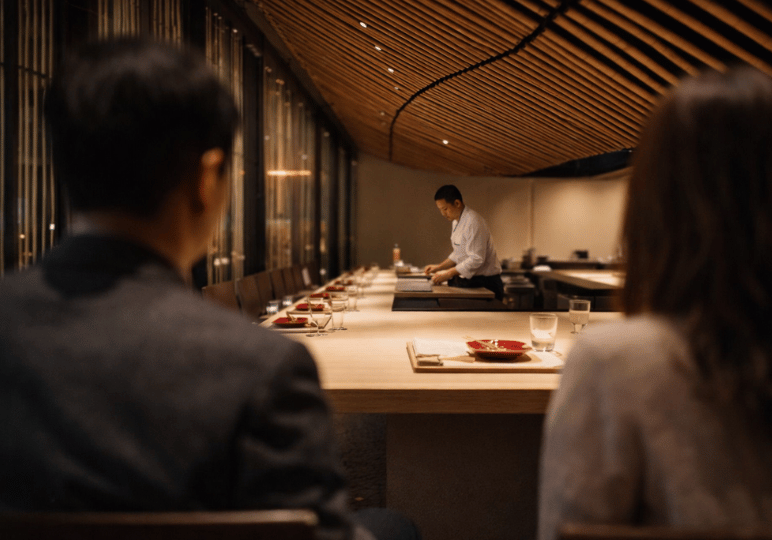 Diners sitting at a sleek, minimalist wooden omakase counter while a chef prepares a meal in the background under warm, elegant lighting.