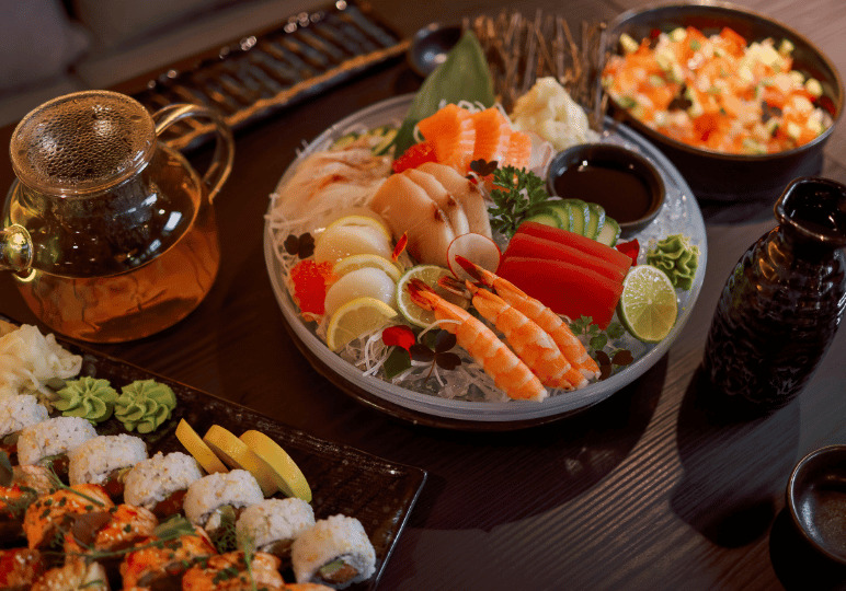 A lavish, fresh sashimi platter served over ice alongside a teapot, assorted sushi rolls, and side dishes on a dark wooden dining table.