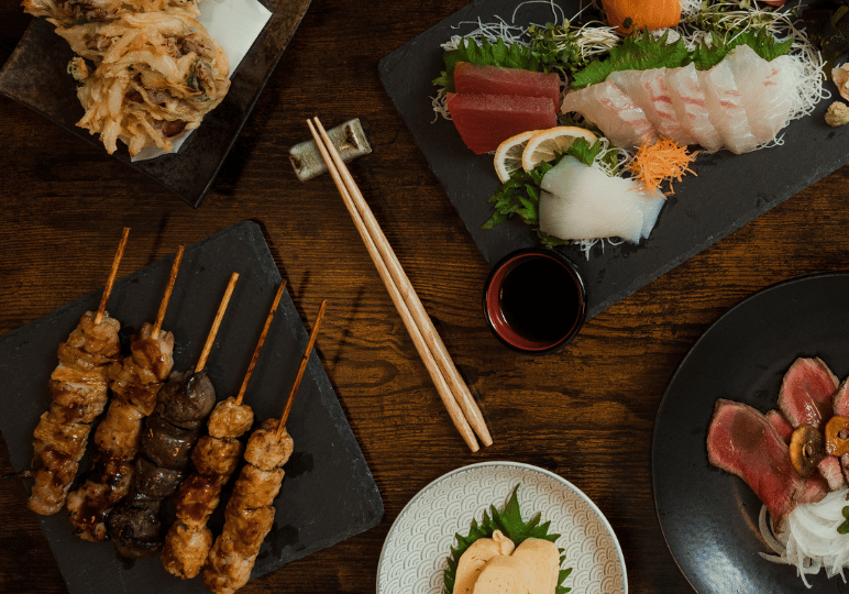 An overhead view of a rustic wooden table filled with diverse Japanese dishes, including yakitori skewers, sashimi, seared beef, tempura, and tamagoyaki.