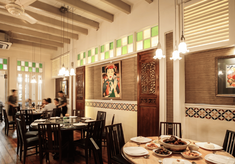 A warmly lit restaurant dining room with heritage-style decor, featuring exposed wooden ceiling beams, green and white stained-glass transoms, and a framed portrait painting on a textured wall. In the foreground, a round wooden table is set for a family-style meal with plates of white rice and several shared dishes. In the background, blurred diners enjoy a communal meal while a server attends to their table, evoking a nostalgic and welcoming atmosphere.