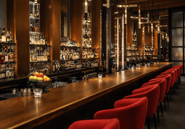 finedining-sg- (5) A sophisticated, warmly lit bar inside a high-end restaurant, showcasing tall illuminated shelves stocked with premium spirits, a long polished wooden counter, and plush red velvet chairs.