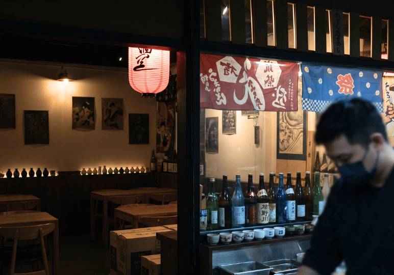 izakaya-afterwork-sg- (9) A dimly lit, traditional izakaya entrance featuring a window display lined with sake bottles, colorful Japanese banners, a glowing red lantern, and a blurred staff member in the foreground.