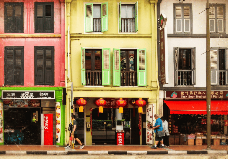 old-chinatown-sg-(1) A row of brightly colored, historic shophouses featuring aging restaurants and traditional shops on the ground floor. The weathered but vibrant facades in deep pink, mustard yellow, and white—complete with open wooden shutters, hanging red lanterns, and classic storefront signage—capture the enduring, nostalgic charm of legacy dining neighborhoods.