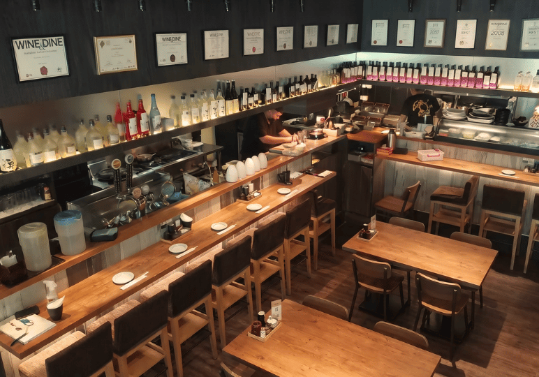 shunjuu-review- (2) Elevated view of the restaurant interior at Shunjuu Izakaya, showcasing the long wooden counter, bar seating, and shelves stocked with sake bottles.