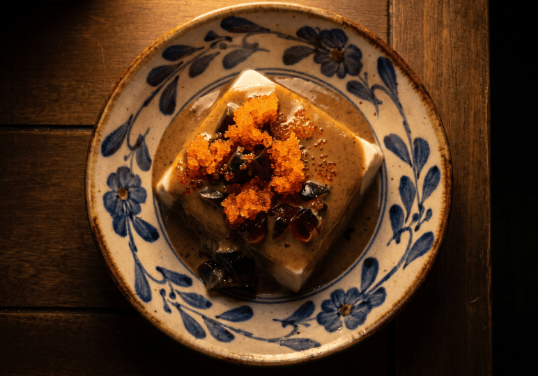 shunjuu-review- (3) A plate of chilled silken tofu, sesame sauce, and bright orange tobiko, served on a blue and white patterned ceramic dish.