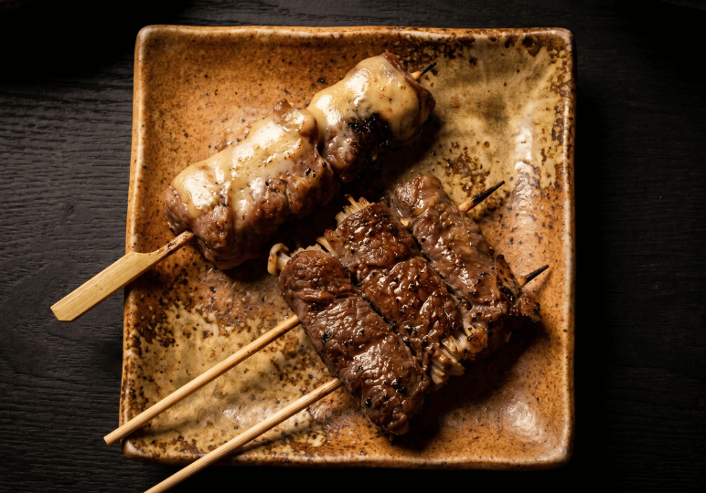shunjuu-review- (4) Close-up of grilled kushiyaki skewers at Shunjuu Izakaya, featuring Scallop Rolled with Pork topped with melted cheese alongside beef-wrapped enoki mushrooms on a rustic square plate.