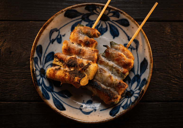 shunjuu-review- (5) Top-down view of two Ton Toro (Pork Cheek) skewers grilled with leeks at Shunjuu Izakaya, showing a slight char and served on a blue-and-white patterned ceramic plate.