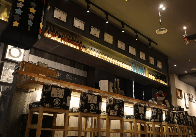 shunjuu-review- (6) A low-angle shot of the bar area at Shunjuu Izakaya featuring high wooden stools with patterned fabric covers and backlit shelves filled with Japanese alcohol.